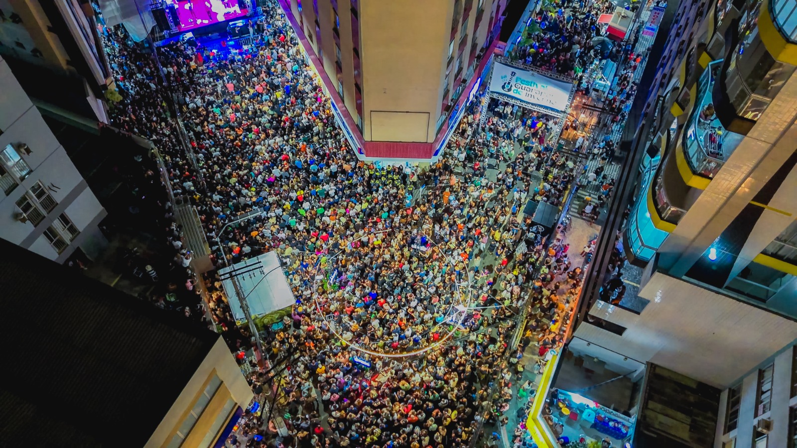 Vista aérea noturna da multidão lotando a Avenida Joaquim da Silva Lima, no Centro de Guarapari, com palco e telão iluminados ao fundo durante o Festival Guarapari de Inverno